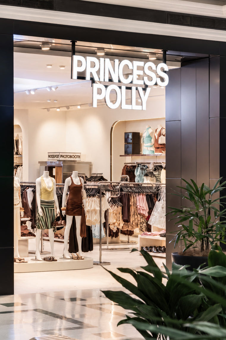Princess Polly clothing store entrance with illuminated white brand signage above a wide opening, showing mannequins and racks of apparel inside.