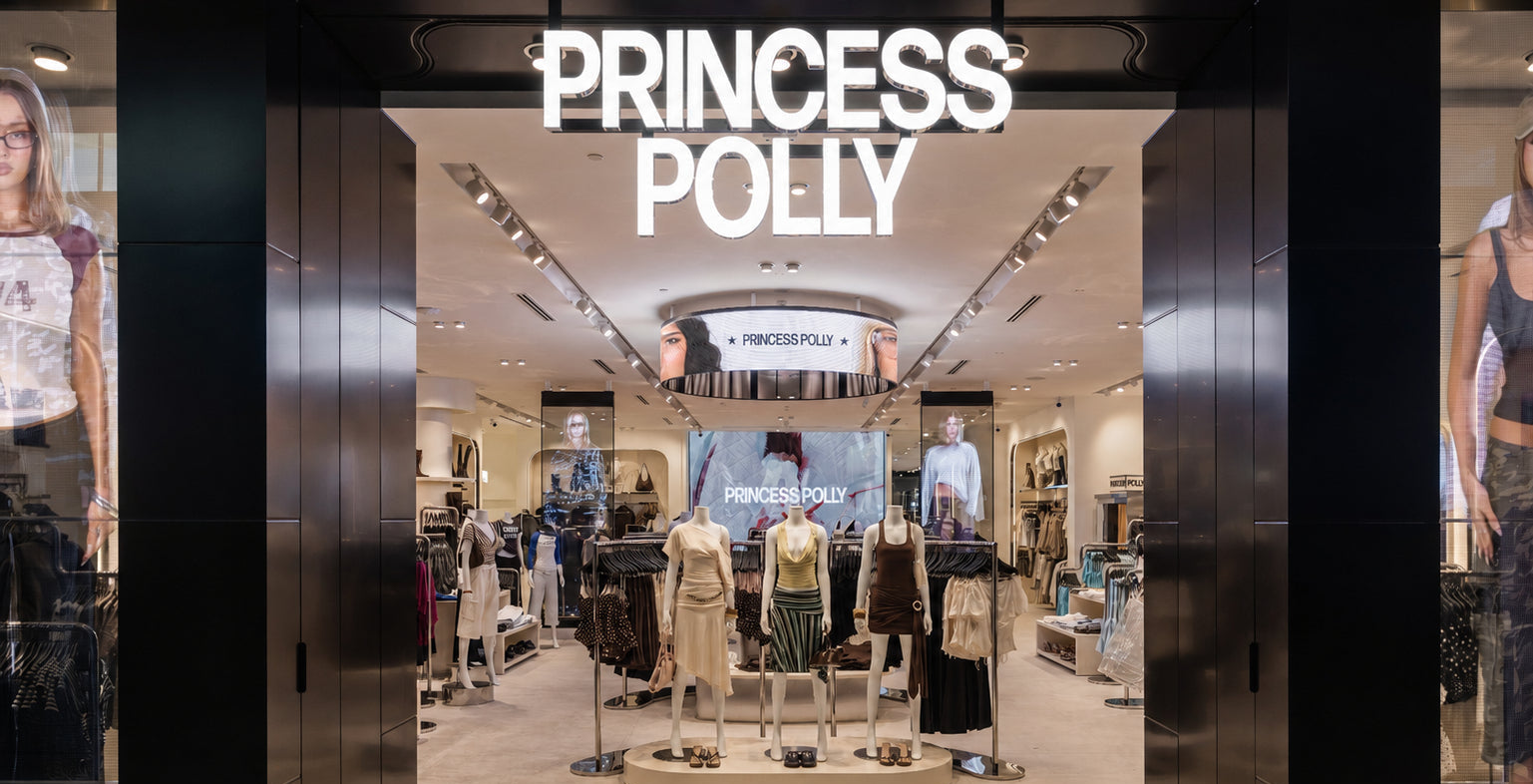Princess Polly clothing store entrance with illuminated white brand signage above a wide opening, showing mannequins and racks of apparel inside.